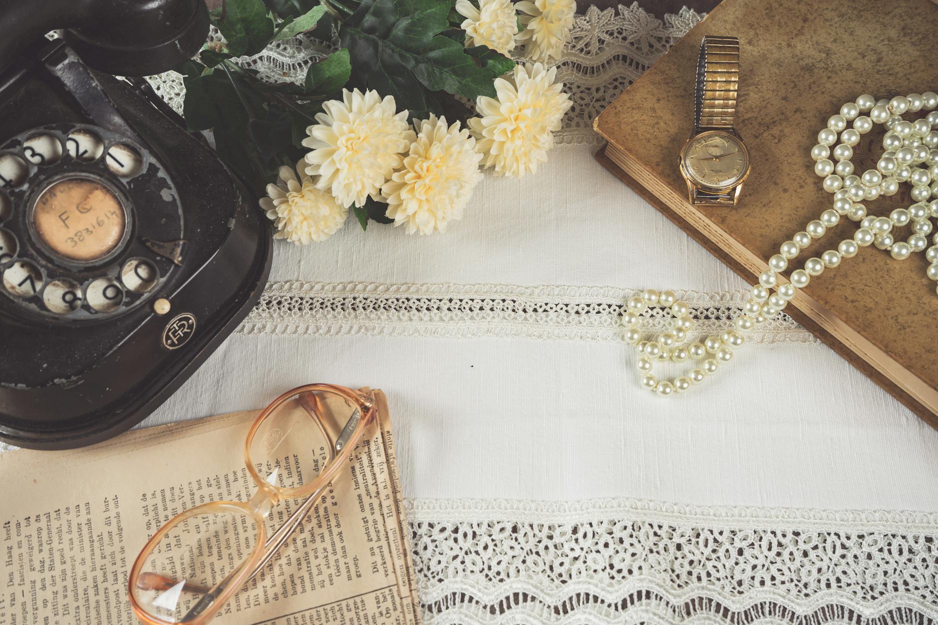 A charming vintage still life featuring an old telephone, pearl necklace, and more on lace cloth.