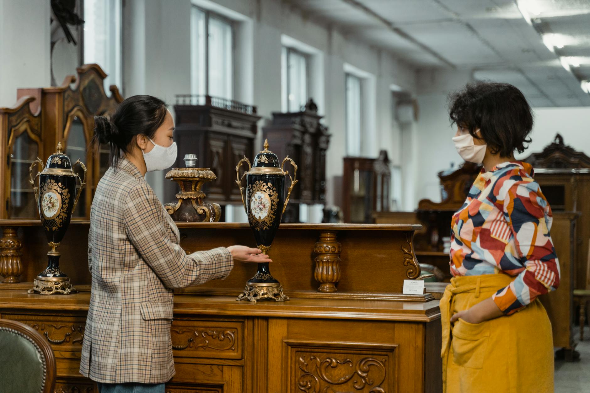 Two women in face masks interacting in an antique store with vintage furniture.
