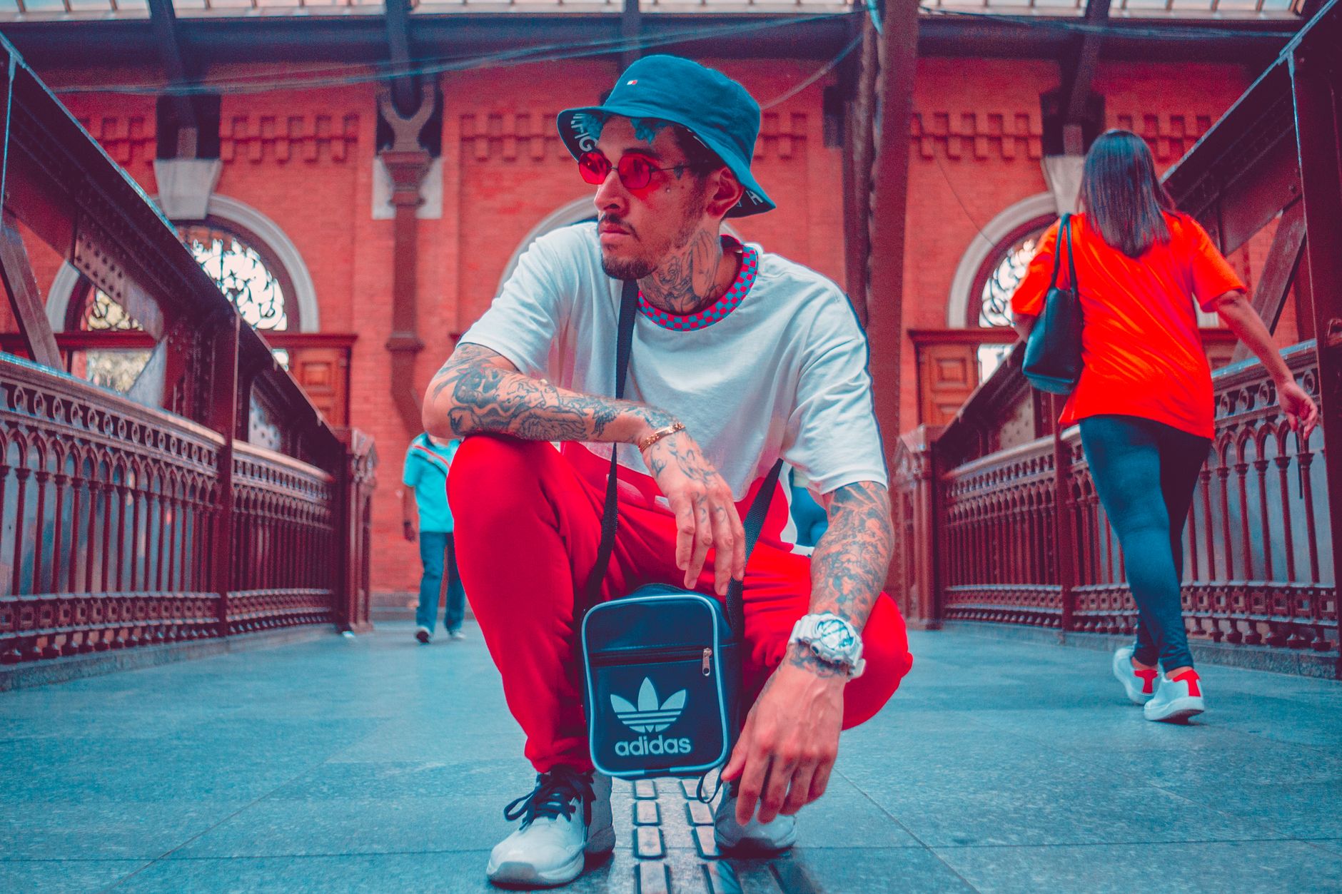 Fashionable tattooed man with sunglasses squatting on a bridge, showcasing urban style.
