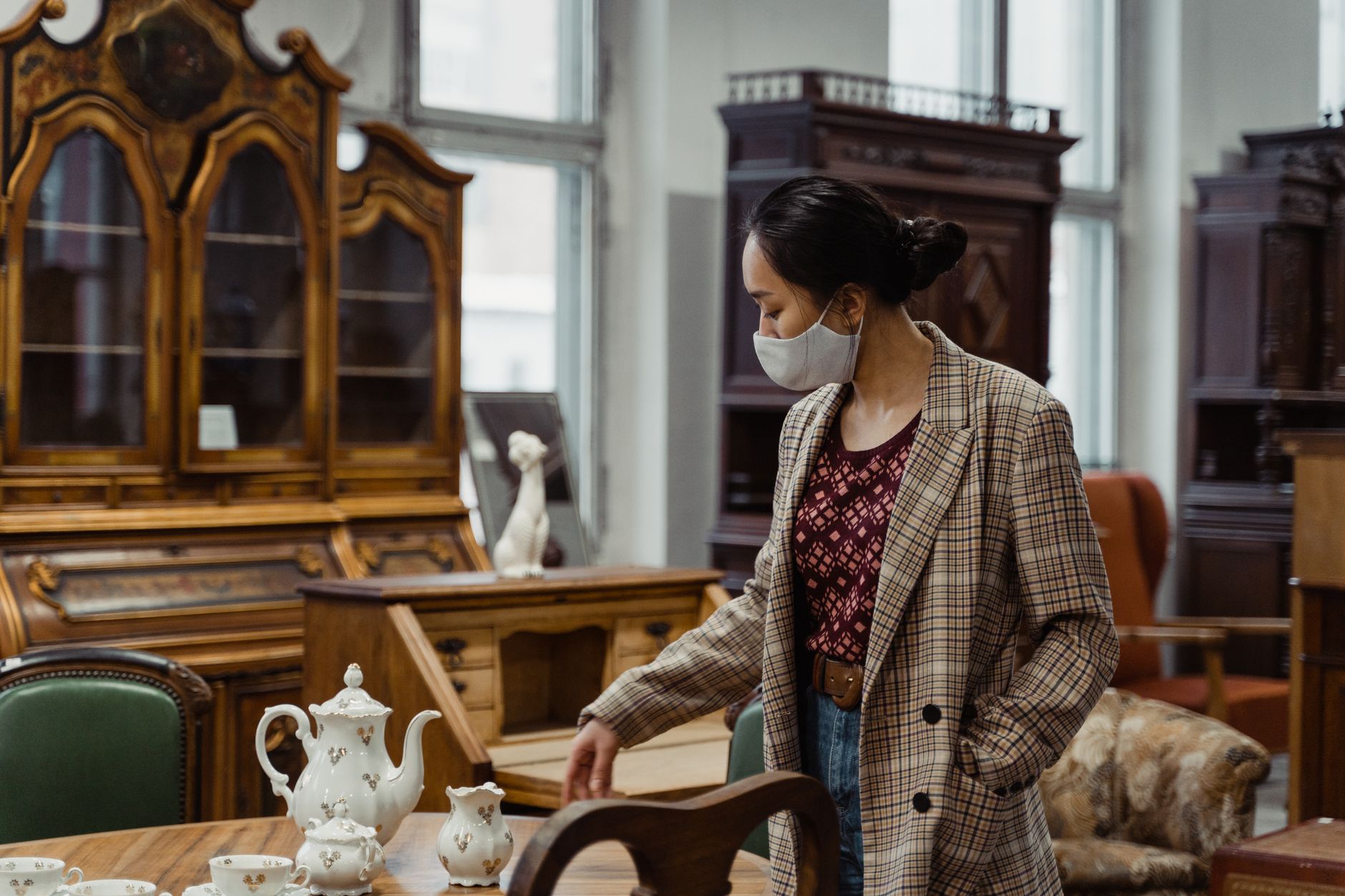 Woman in plaid coat and mask browsing porcelain tea set in antique store.