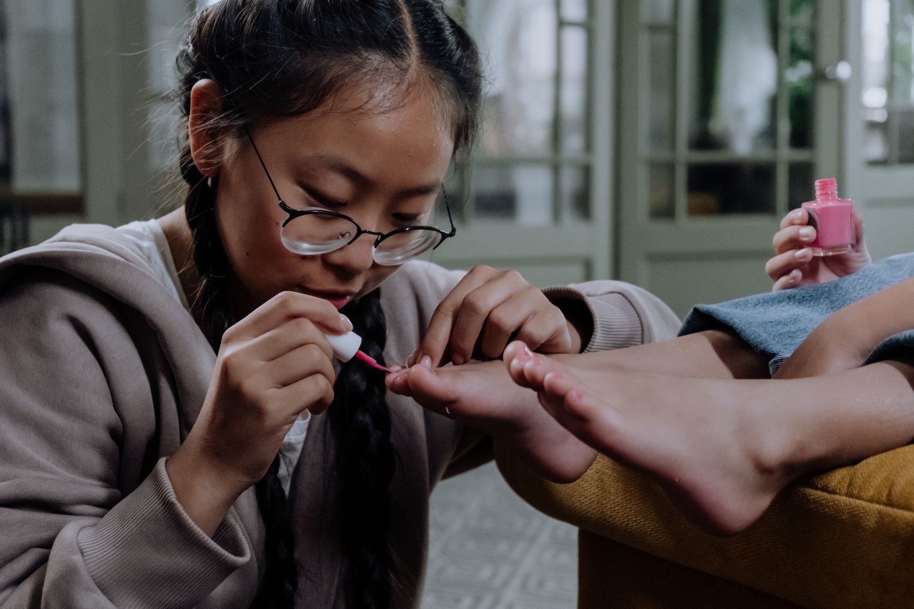 Young girl applying pink nail polish indoors, close-up on pedicure with eyeglasses.